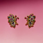 Butterfly Bloom stud earrings featuring pink and blue sapphires, white diamonds and green tsavorite, photographed against a pink background