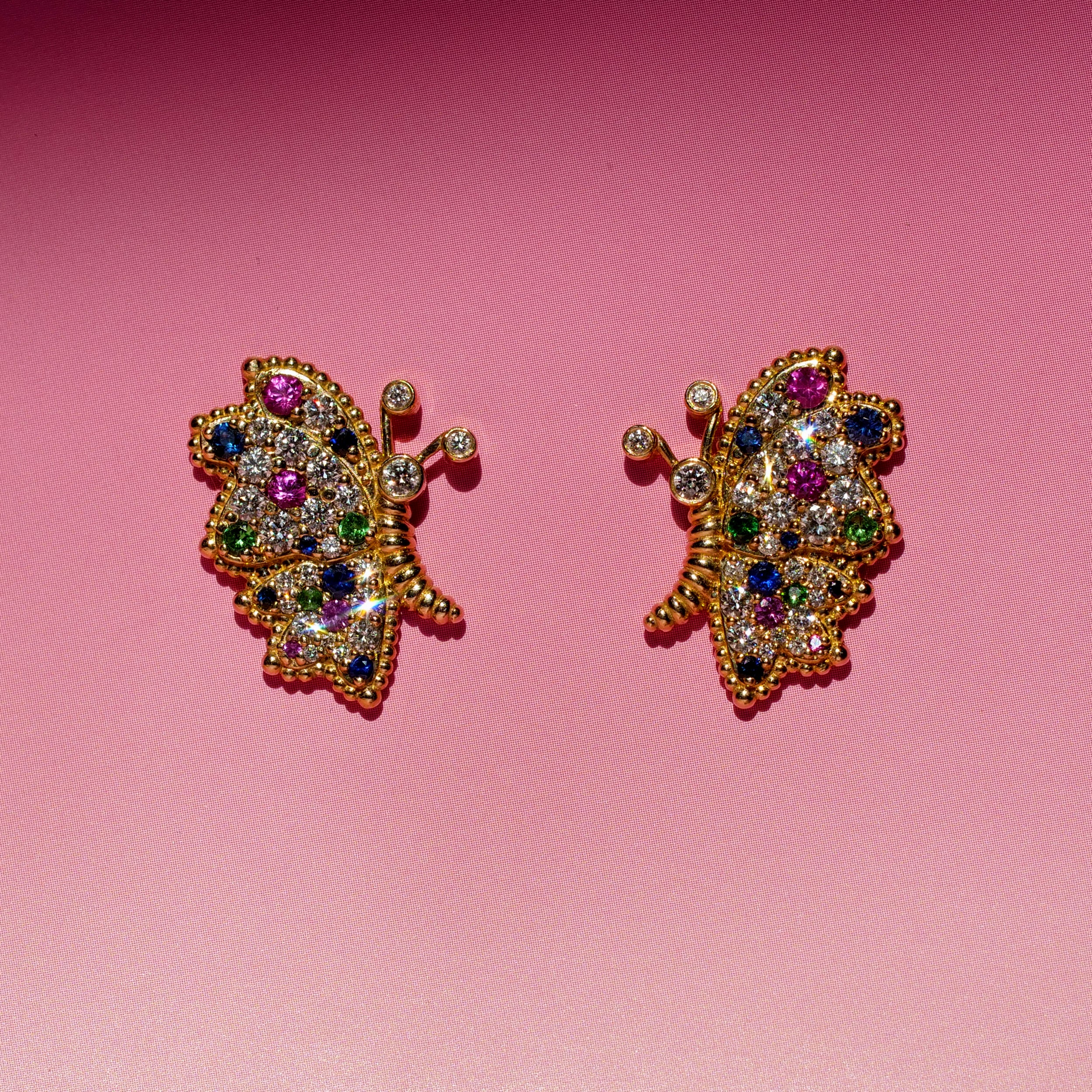 Butterfly Bloom stud earrings featuring pink and blue sapphires, white diamonds and green tsavorite, photographed against a pink background